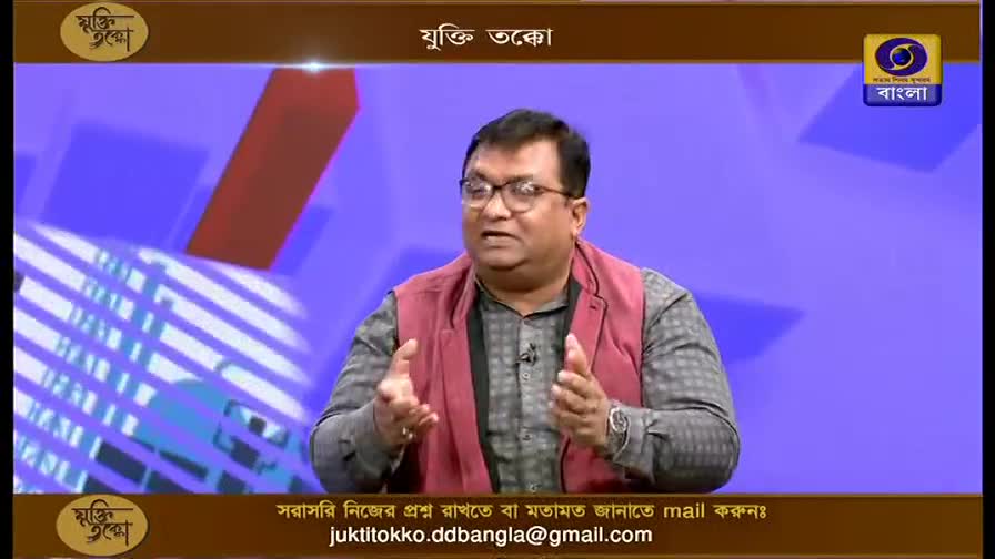 A man with glasses gestures with both hands while speaking on a DD Bangla program. The show's title, "Yukti Takko," appears above him.
