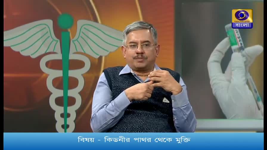 A man in a blue shirt and dark vest gestures with his hands on a set for DD Bangla. To his right, a gloved hand holds a syringe.