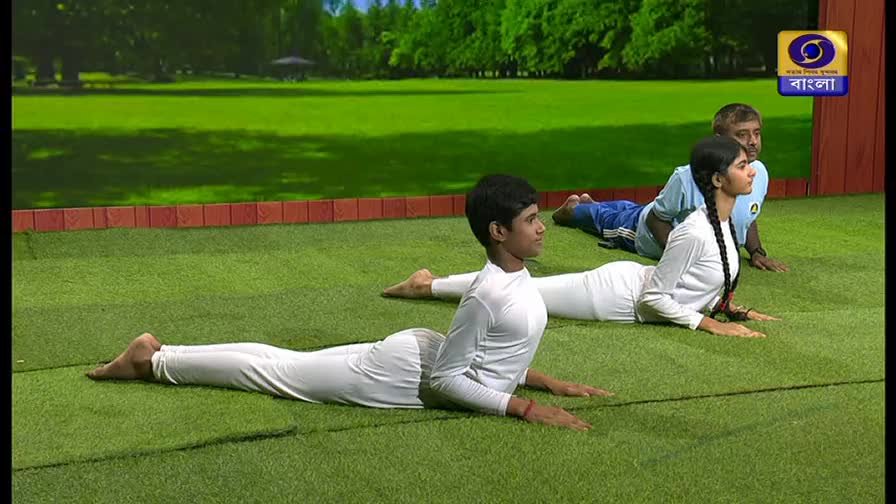 Three people in white practice yoga poses on a green mat. A boy in the foreground arches his back, while a girl behind him mirrors the pose. A man in a blue shirt lies further back, also in a similar position.