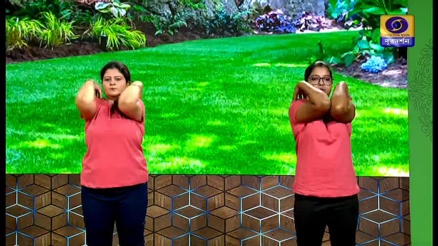 Two women stand on a stage, each with their arms crossed and hands resting on the opposite shoulder. A vibrant green backdrop, reminiscent of a park, stretches behind them, with the DD Bangla logo visible in the corner.