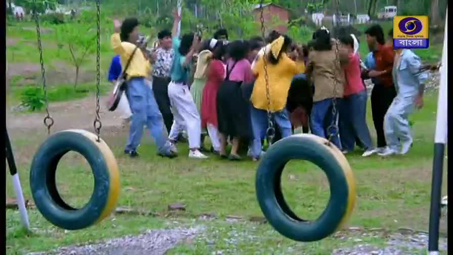 A group of young people are dancing in a circle on the grass, their arms raised. A tire swing sways gently in the foreground, its yellow and green painted surface catching the light.