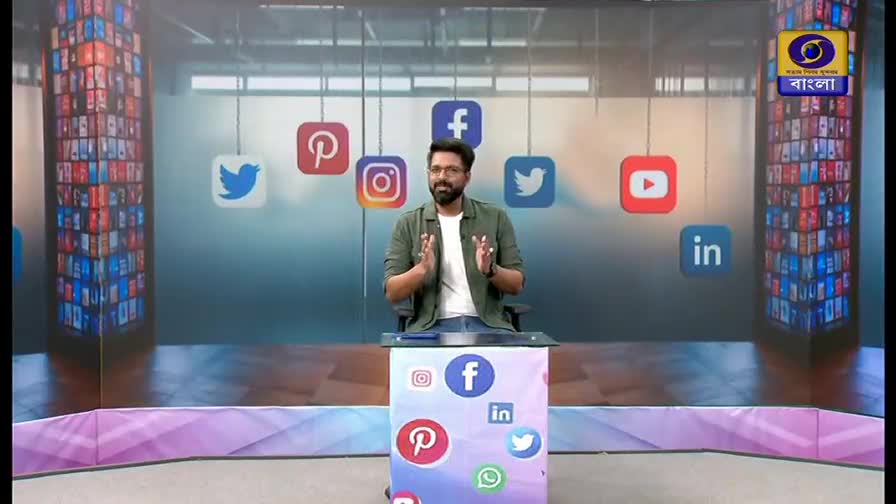 A man stands at a podium, his hands clasped in front of him, as social media logos float in the background. The DD Bangla logo is visible in the upper right corner, hinting at a broadcast from India.