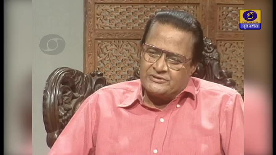 A man with glasses, wearing a pink shirt, sits in a carved wooden chair. The DD Bangla logo is visible in the corner of the screen.