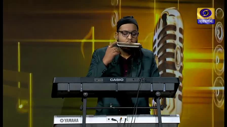 A man plays a harmonica and a keyboard on a stage, likely for a DD Bangla broadcast. The backdrop features a stylized microphone and the Doordarshan logo.