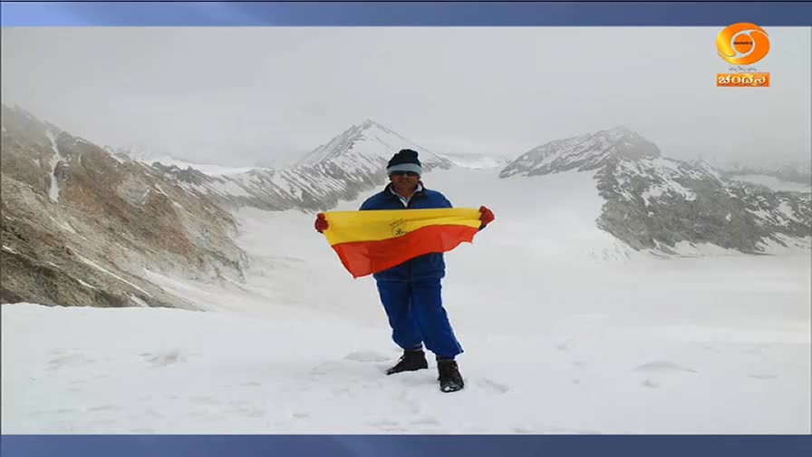 A man stands in the snow, holding a flag with red and yellow stripes against a backdrop of snow-covered mountains. The DD Chandana logo is visible in the corner, suggesting this is part of a broadcast from India.
