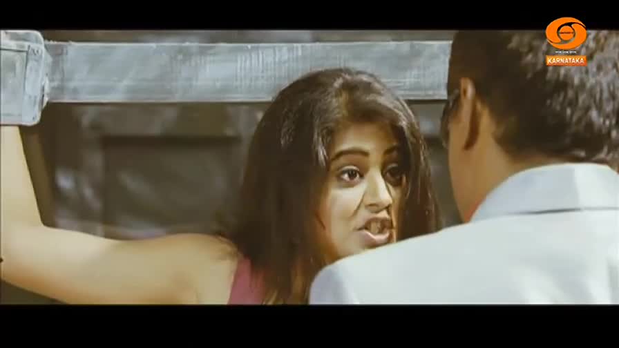 A woman is leaning against a metal bar, her expression intense as she speaks to a man in a white shirt. The scene is set in what appears to be a studio, with the DD Chandana logo visible in the corner.
