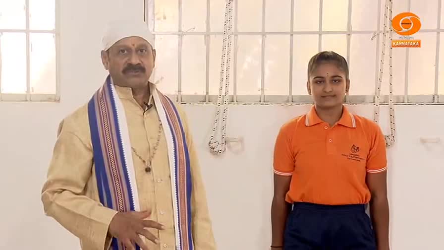 A man in a tan kurta and a colorful shawl stands with his hand on his stomach, while a young woman in an orange polo shirt and dark shorts stands beside him. The DD Chandana logo is visible in the corner, indicating this is a broadcast from Karnataka, India.