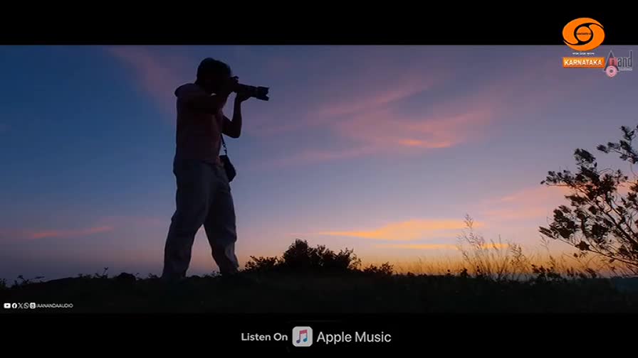 A lone figure stands silhouetted against the twilight sky, raising a camera to capture the fading light. The colors of the sunset, a blend of soft pinks and oranges, stretch across the horizon, painting the sky above the Indian landscape.