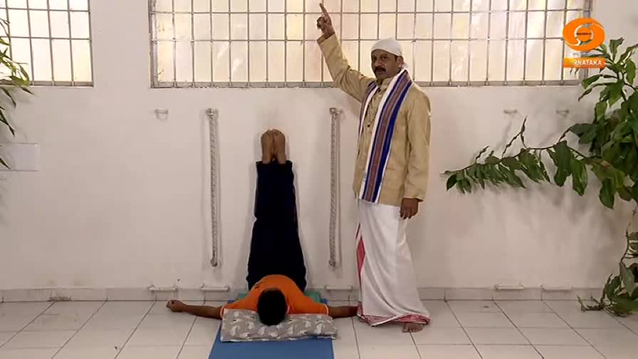 A man in a white dhoti and a tan jacket, with a colorful sash, stands to the right, pointing upwards. To his left, another man lies on a mat, his legs and torso raised vertically, supported by his shoulders. This is likely a segment from DD Chandana.