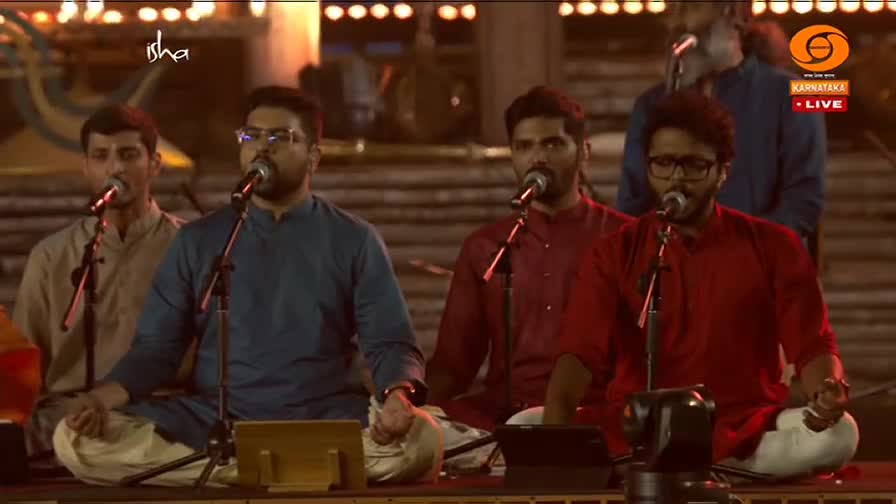 Four men in traditional Indian attire sit cross-legged, singing into microphones on a stage. The DD Chandana logo is visible, indicating a broadcast from Karnataka.