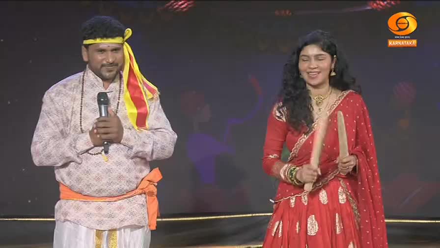 A man in traditional attire holds a microphone, while a woman in a red sari beats two wooden sticks together. They stand on a stage, likely for a performance broadcast on DD Chandana.