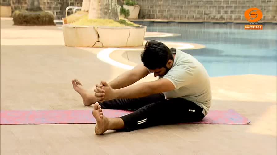 A man in a light green t-shirt and black track pants bends forward, hands clasped, his legs extended on a pink yoga mat. This stretch, part of a DD Chandana program, takes place outdoors near a pool.