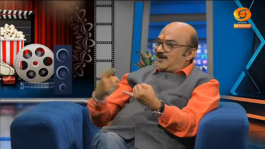 A man in an orange shirt and grey vest gestures animatedly while speaking. Behind him, a backdrop features film reels, popcorn, and the DD Chandana logo.