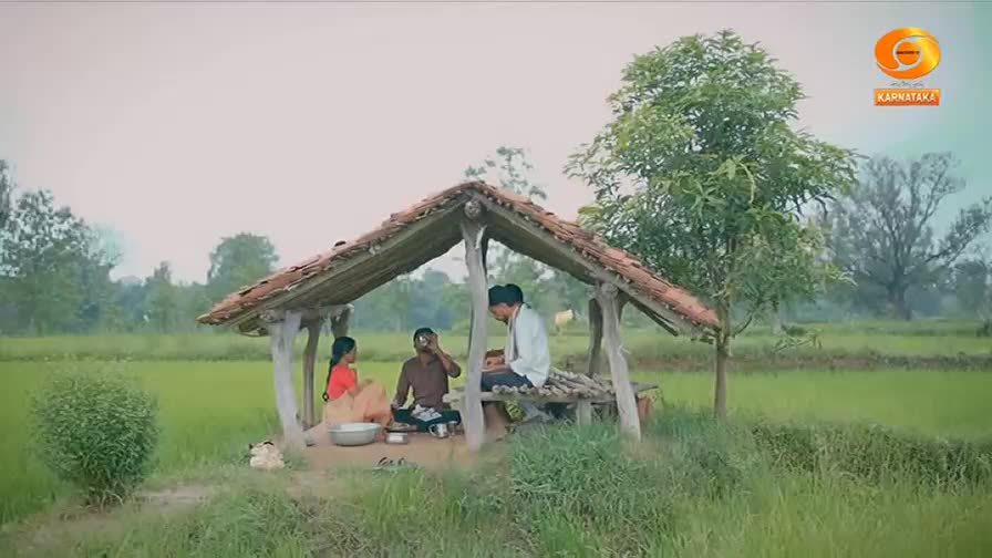 A young woman in a bright orange sari sits with a man in a brown shirt, sharing a meal under a thatched roof shelter. Another man, dressed in white, joins them, his attention focused on something in his hands. The scene unfolds amidst lush green paddy fields, a typical backdrop for rural Karnataka.