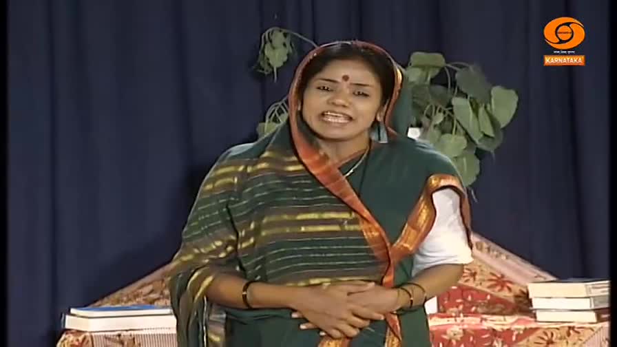 A woman in a green sari with an orange border speaks animatedly, her hands clasped at her waist. Behind her, a dark blue curtain forms the backdrop, with a few leafy branches visible to her right.