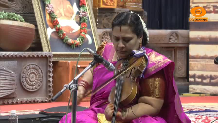 A woman in a vibrant pink sari plays the violin, her bow moving across the strings. Behind her, a framed portrait adorned with a floral garland hangs on the wall. A woman in a vibrant pink sari plays the violin, her bow moving across the strings. Behind her, a framed portrait adorned with a floral garland hangs on the wall.
