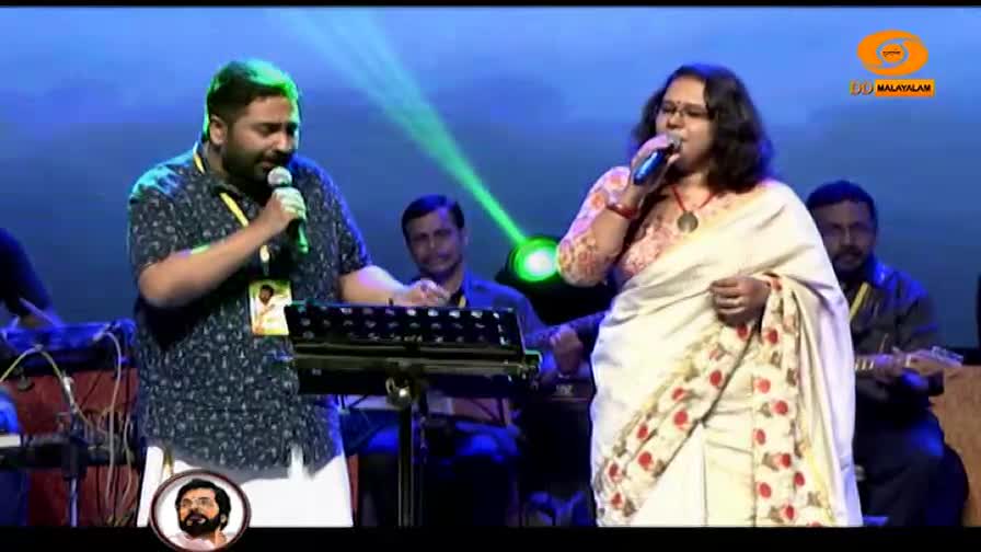 A man in a patterned shirt sings into a microphone, his gaze fixed on the keyboard in front of him. Beside him, a woman in a white sari belts out a tune, her eyes closed in concentration. A man in a patterned shirt sings into a microphone, his gaze fixed on the keyboard in front of him. Beside him, a woman in a white sari belts out a tune, her eyes closed in concentration.