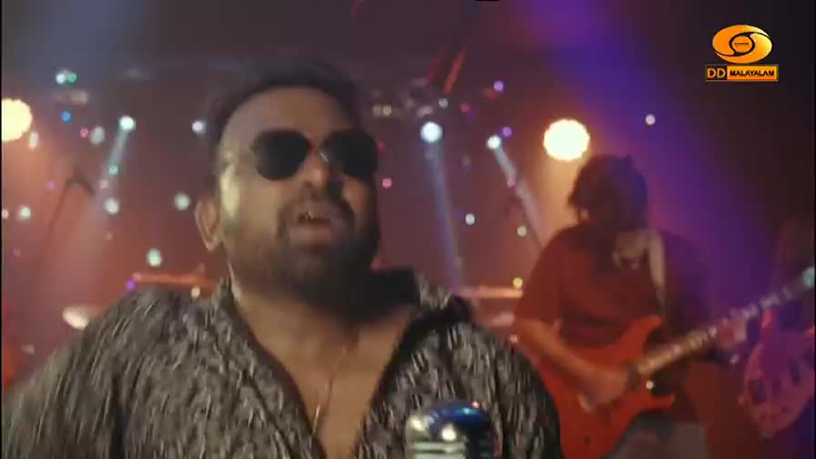 The lead singer, wearing dark sunglasses, belts into a microphone on a stage lit by colorful lights. Behind him, a guitarist in a red shirt plays an electric guitar. The lead singer, wearing dark sunglasses, belts into a microphone on a stage lit by colorful lights. Behind him, a guitarist in a red shirt plays an electric guitar.