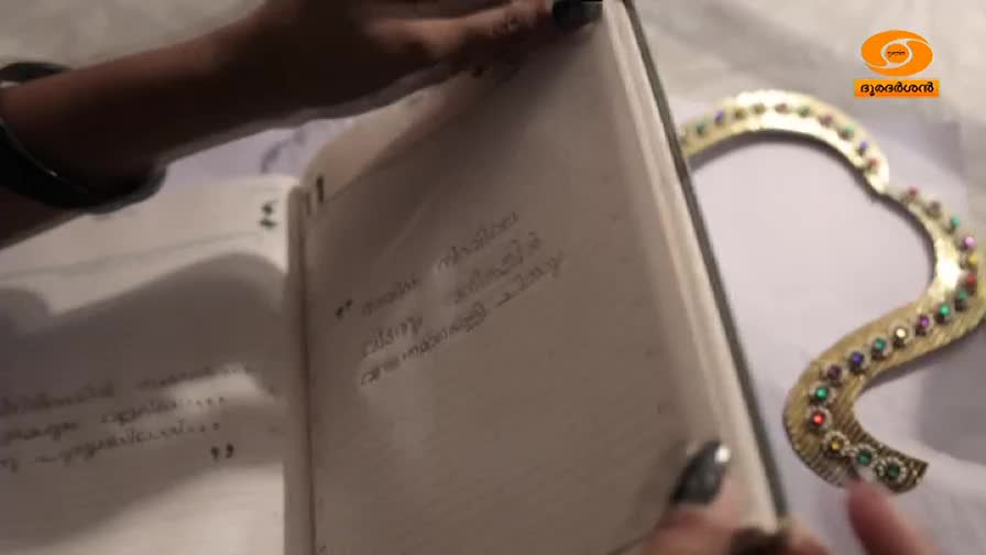 Fingers trace the Malayalam script in an open notebook. A gilded, jeweled heart rests beside it. Fingers trace the Malayalam script in an open notebook. A gilded, jeweled heart rests beside it.