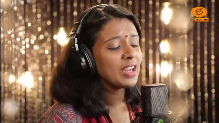 A woman with headphones on sings into a microphone, her eyes closed in concentration. The DD Malayalam logo is visible in the background. A woman with headphones on sings into a microphone, her eyes closed in concentration. The DD Malayalam logo is visible in the background.