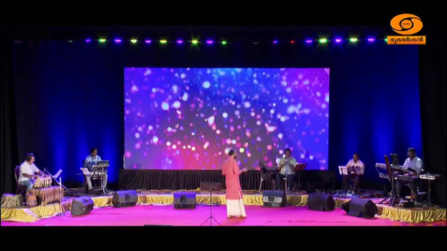 A singer in a peach-colored kurta stands center stage, facing a band of musicians. To his left, a man plays the tabla, while to his right, two men play keyboards, all backed by a vibrant, abstract light display on a large screen. This performance is being broadcast by DD Malayalam. A singer in a peach-colored kurta stands center stage, facing a band of musicians. To his left, a man plays the tabla, while to his right, two men play keyboards, all backed by a vibrant, abstract light display on a large screen. This performance is being broadcast by DD Malayalam.