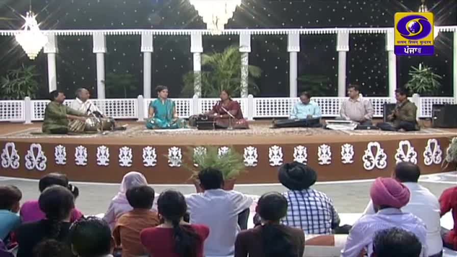 A group of musicians sits on a raised stage, performing for an audience. A woman in a teal outfit plays a harmonium while a man beside her taps a tabla, their music broadcast on DD Punjabi.