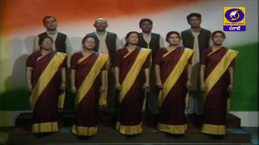 A group of people stand before a backdrop resembling the Indian flag. Three women in maroon saris with yellow borders stand in the front row, while four men in vests over shirts are positioned behind them. The DD Punjabi logo is visible in the upper right corner.