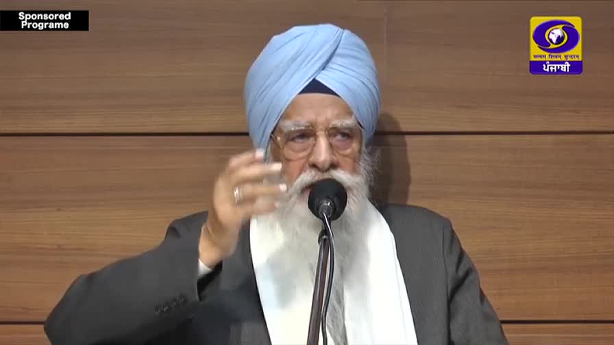 An elder man with a long white beard and a light blue turban speaks into a microphone. He gestures with his right hand, his fingers splayed.