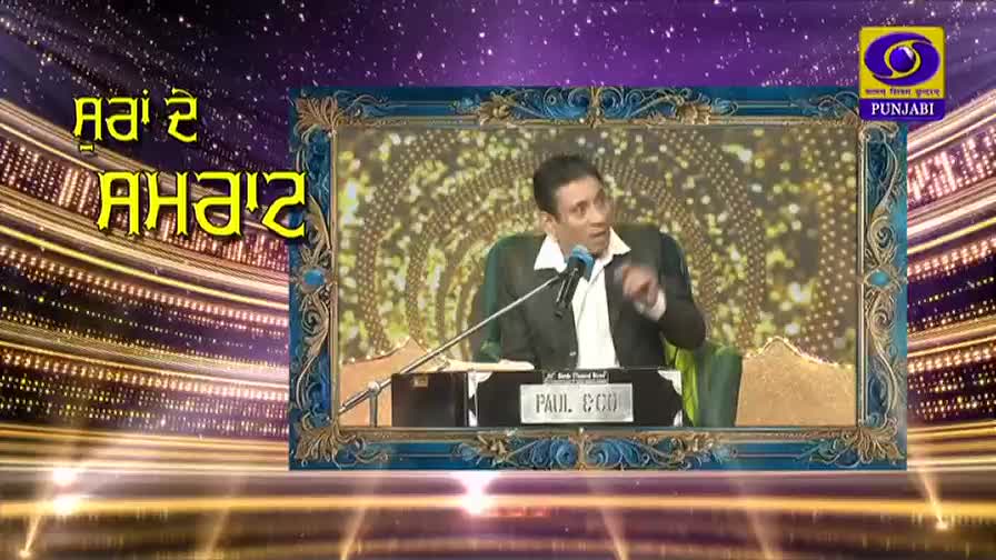 A man in a dark suit and white shirt gestures with his right hand while speaking into a microphone. Behind him, a colorful, ornate frame encloses a glittering backdrop, and the DD Punjabi logo is visible in the upper right corner.