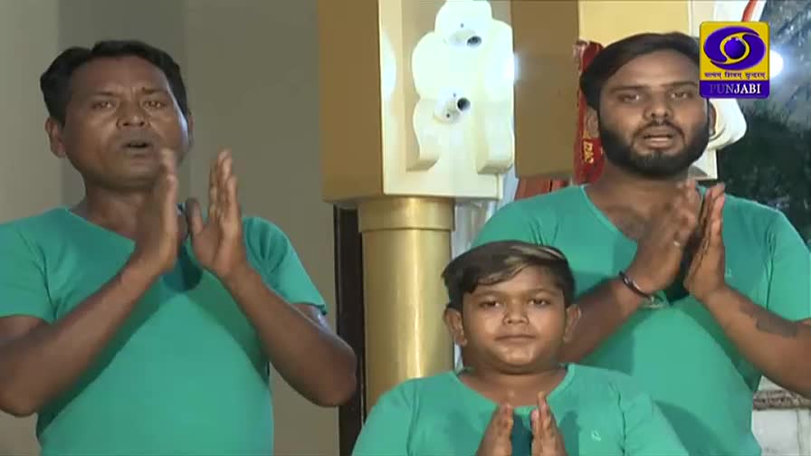 Three men in teal shirts stand with their hands pressed together in front of them. The man on the left is speaking, his mouth open wide. A logo for DD Punjabi appears in the upper right corner.