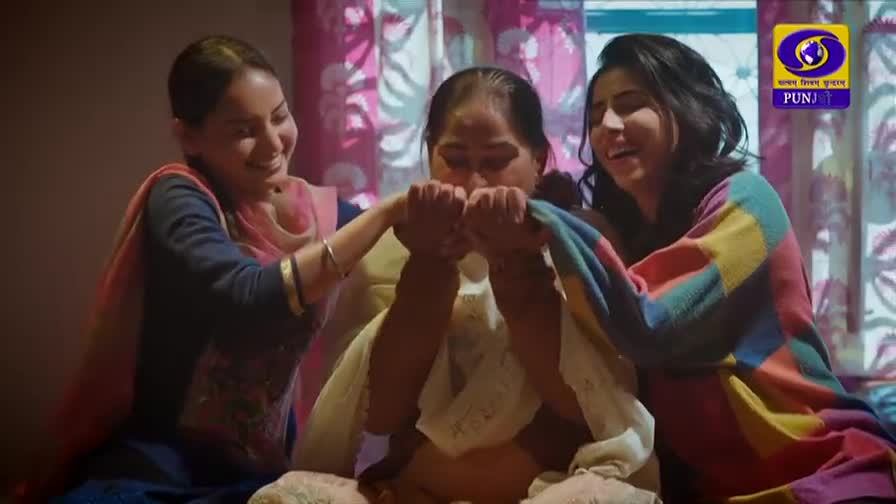 Two young women playfully pull a colorful shawl over their mother's face. Their laughter fills the room, a scene likely from a DD Punjabi program. Two young women playfully pull a colorful shawl over their mother's face. Their laughter fills the room, a scene likely from a DD Punjabi program.