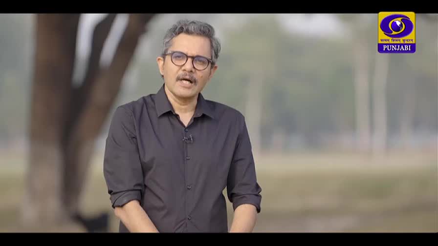 A man in a dark shirt speaks directly to the camera, his glasses reflecting the soft light. Behind him, the DD Punjabi logo is clearly visible. A man in a dark shirt speaks directly to the camera, his glasses reflecting the soft light. Behind him, the DD Punjabi logo is clearly visible.
