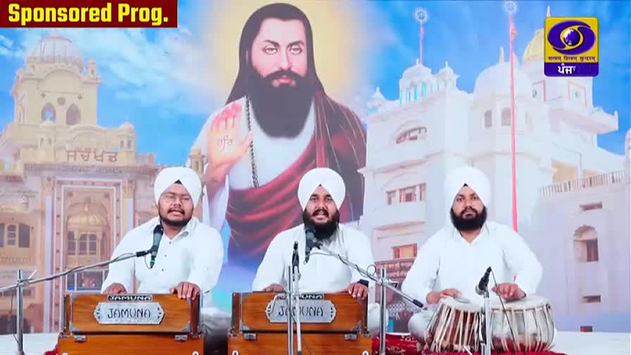 Three men in white turbans and kurtas are seated, each with a musical instrument. The man on the left plays a harmonium, the middle man sings into a microphone, and the man on the right plays a tabla. Behind them, a large portrait of a bearded man with a benevolent expression dominates the backdrop, with a stylized image of a Sikh temple and the DD Punjabi logo. Three men in white turbans and kurtas are seated, each with a musical instrument. The man on the left plays a harmonium, the middle man sings into a microphone, and the man on the right plays a tabla. Behind them, a large portrait of a bearded man with a benevolent expression dominates the backdrop, with a stylized image of a Sikh temple and the DD Punjabi logo.