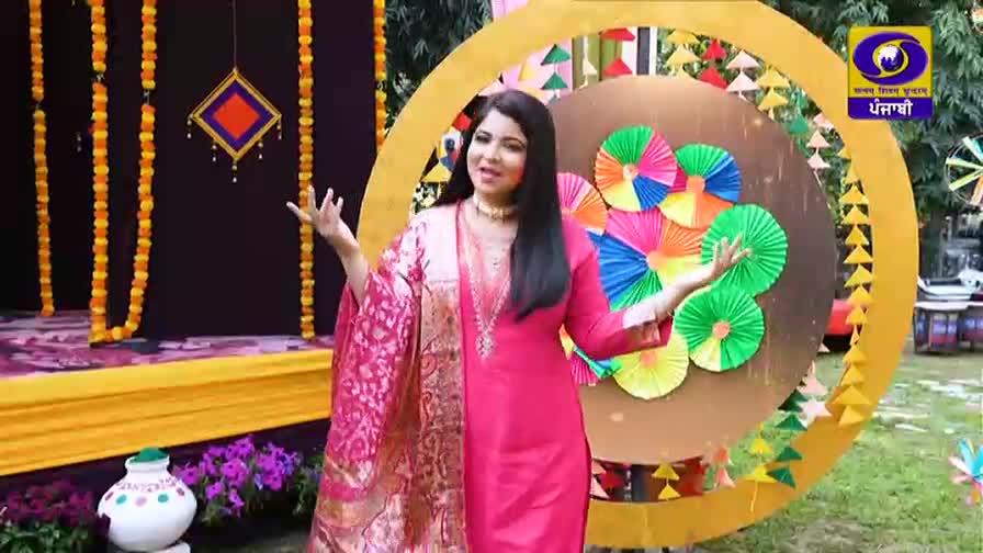 A woman in a vibrant pink suit stands on a decorated stage, her hands gesturing outward. Behind her, colorful paper fans are arranged on a circular backdrop, with the DD Punjabi logo visible. A woman in a vibrant pink suit stands on a decorated stage, her hands gesturing outward. Behind her, colorful paper fans are arranged on a circular backdrop, with the DD Punjabi logo visible.