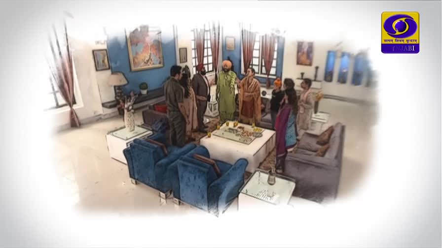 A group of people stand gathered around a low table in a well-furnished room. A man in a green kurta and turban gestures as he speaks to the others.