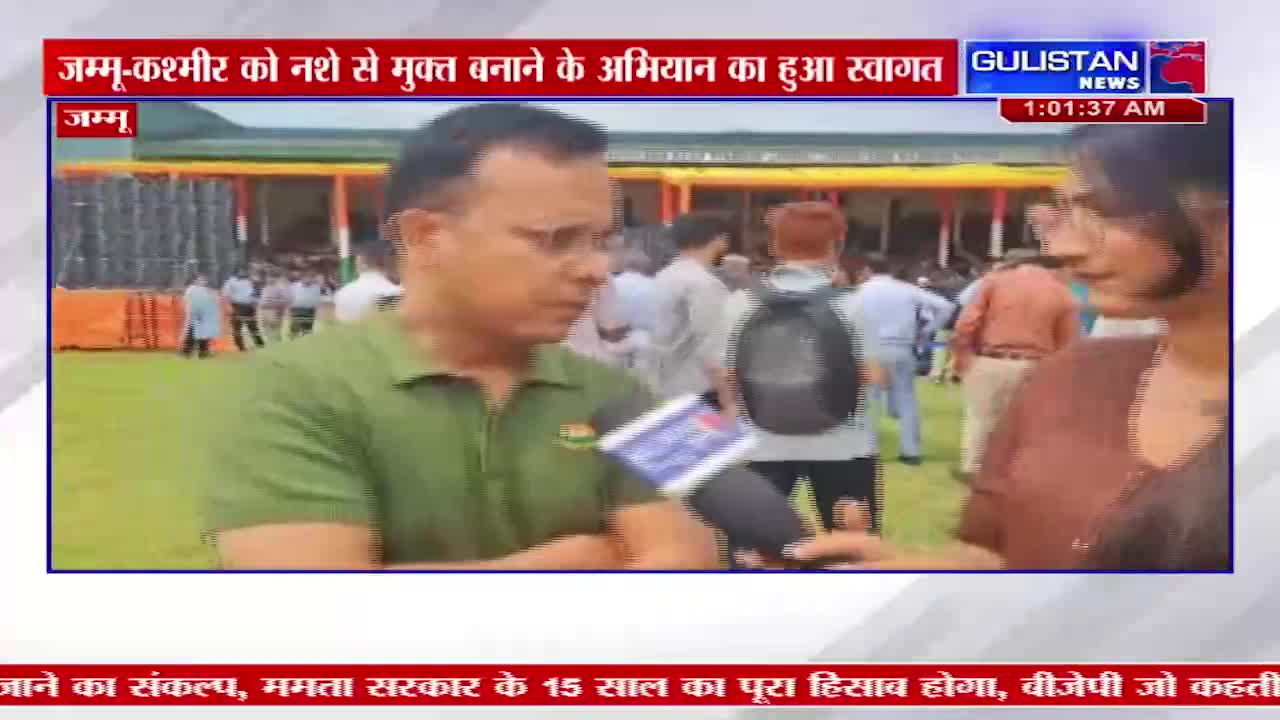 A man in a green shirt is speaking into a microphone, likely for Gulistan News. Behind him, a crowd of people mill about in front of a tented structure.