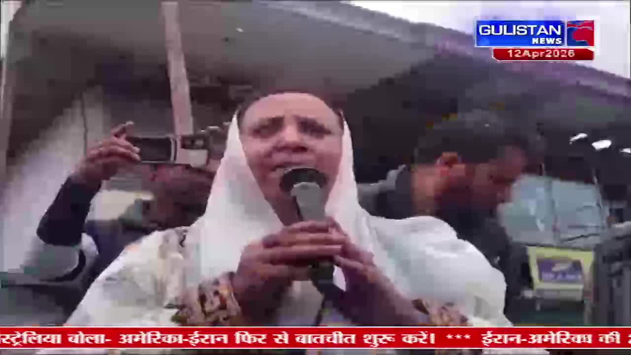 A woman in a white headscarf speaks into a microphone, her face animated. Beside her, a man holds up a phone, recording the scene.