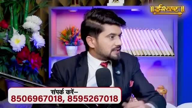 A man in a suit speaks into a microphone, with a colorful floral arrangement to his left. Behind him, a shelf holds rows of books, and the Ishwar logo shines in the corner.