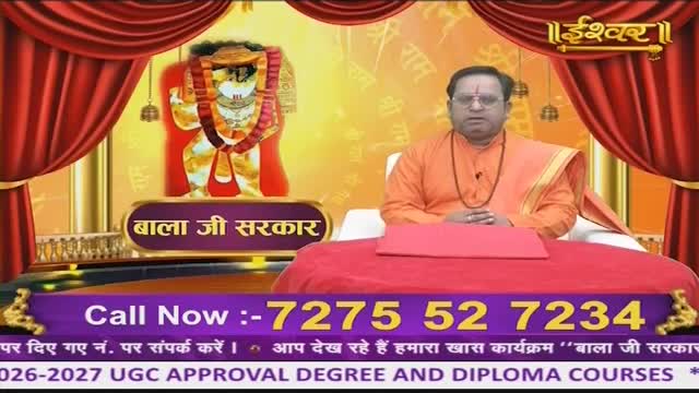 A man in an orange robe sits behind a red table, speaking into a microphone. Behind him, a vibrant image of a deity is displayed, framed by red curtains.