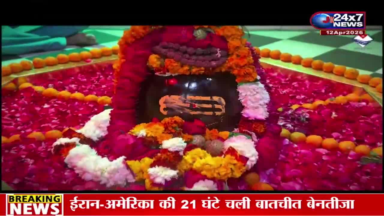 A large, dark Shiva lingam is adorned with vibrant red and pink rose petals, marigold garlands, and white flowers. The offering surrounds the sacred stone, with a glimpse of a person in a blue garment to the left.