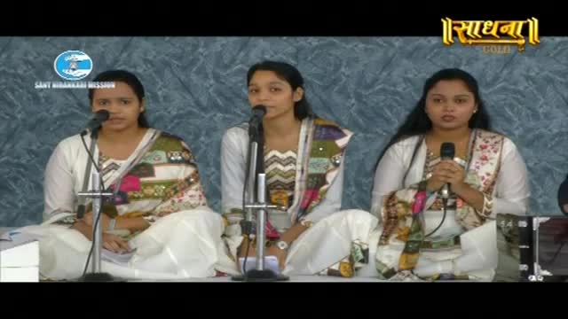 Three women sit on a stage, each with a microphone before them. They wear white kurtis and vibrant dupattas, their faces turned towards the audience. A logo for "Sadhna Gold" is visible in the upper right corner.