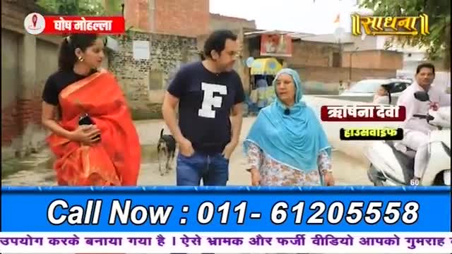 A woman in a bright orange sari walks past a man in a dark t-shirt, while a dog trots behind them. A woman in a blue headscarf stands nearby, and a man on a scooter passes in the background.