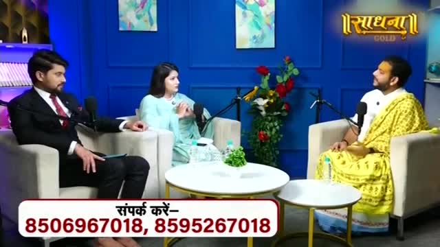 A man in a suit and a woman in a light blue outfit sit facing a man draped in a yellow cloth. The Sadhna Gold logo glows above them.