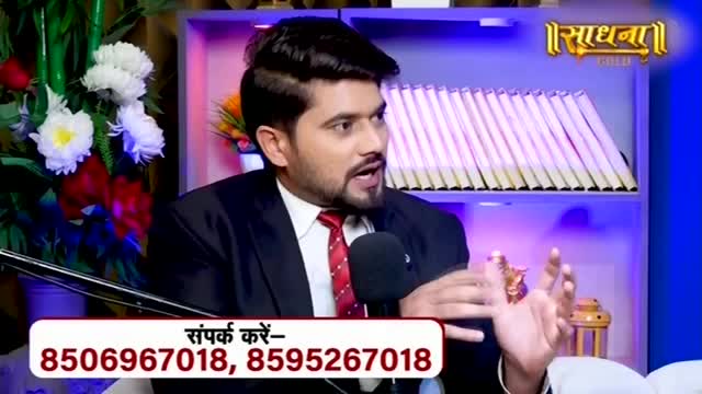 A man in a suit speaks animatedly, his hands gesturing. Behind him, a shelf holds rows of books, and a logo for "Sadhna" glows.