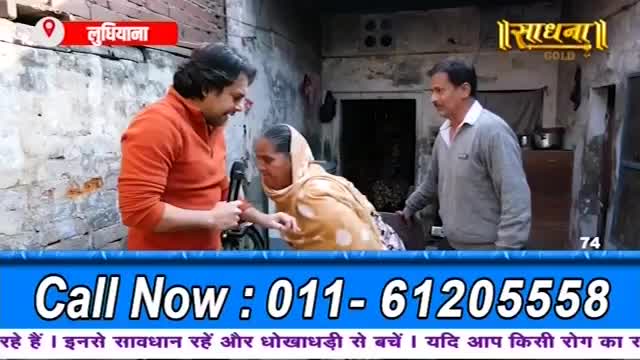 A man in an orange sweater holds a microphone to a woman in a light brown shawl. Another man stands behind her, his hand on her shoulder. The scene appears to be in a courtyard in India.