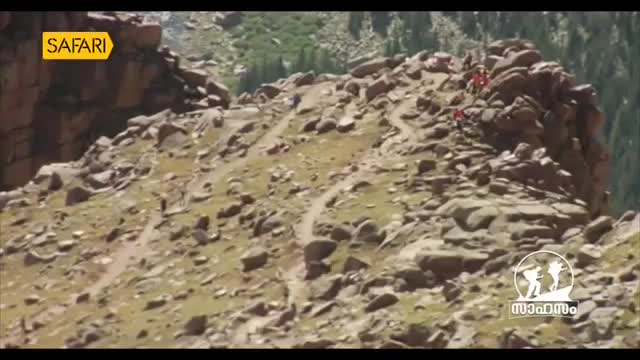 A group of people in red shirts are carefully making their way up a narrow, rocky mountain path. The terrain is steep and uneven, with sparse vegetation clinging to the slopes.