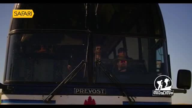 A large bus, marked with "Safari" and a Malayalam logo, approaches. The driver, visible through the windshield, looks focused on the road.