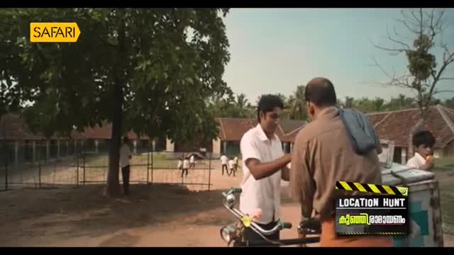 Two men stand near a bicycle, one in a white shirt, the other in a brown one. A young boy watches from the side, with a building and trees in the background.