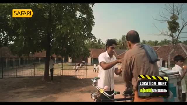 A young man in a white shirt offers something to an older man, his hand outstretched. Behind them, a school building and a few cows are visible under a bright sky.