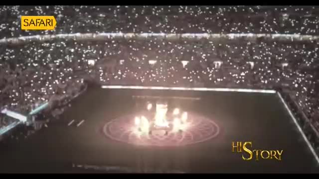 A ring of performers moves in unison on a circular stage, illuminated by a bright light. The stadium crowd holds up thousands of small lights, creating a dazzling spectacle.